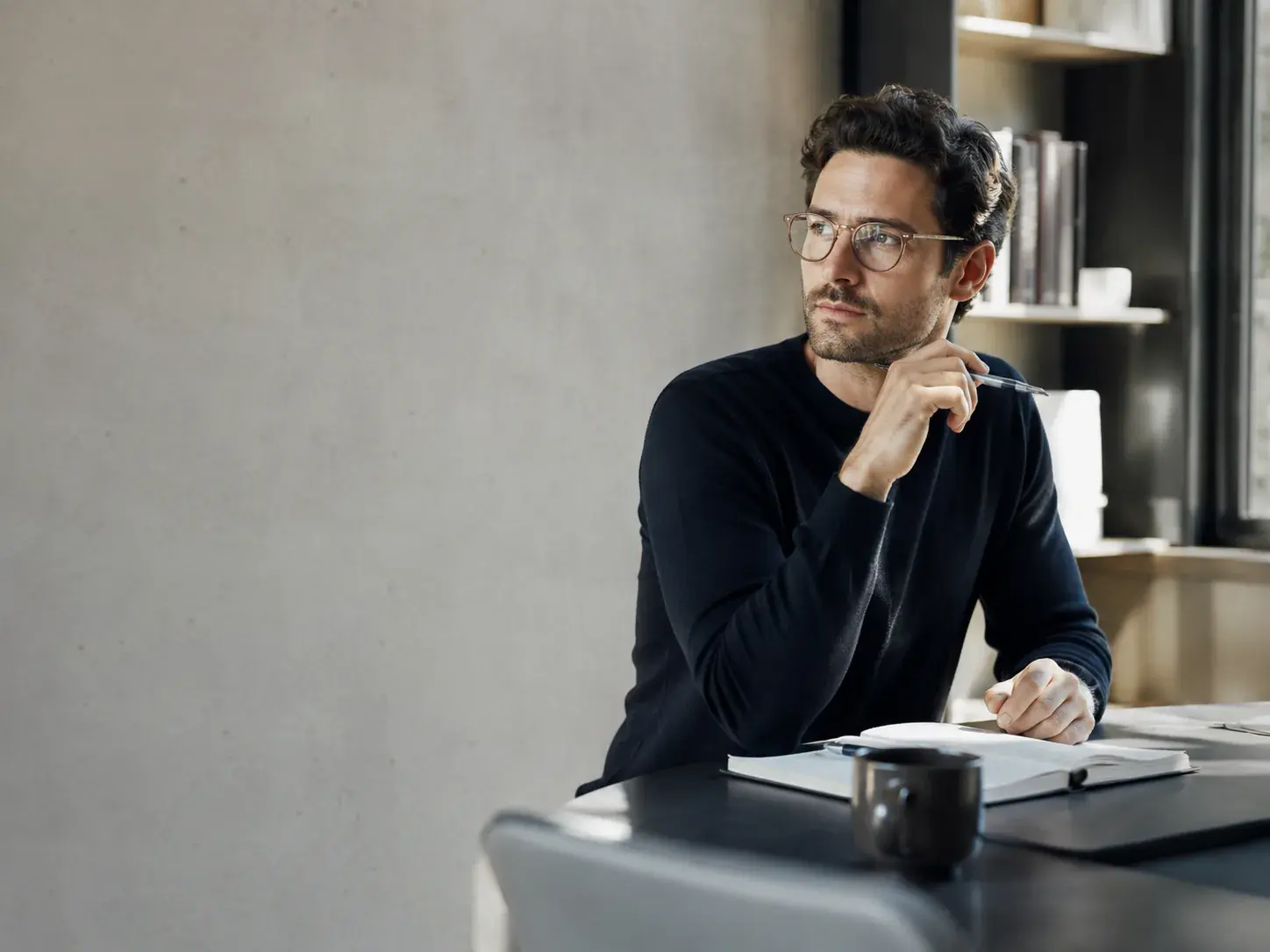 A person in a dark jumper sits at a desk in a bright room, holding a pen and looking away in thought, with an open notebook and a mug in front of them.